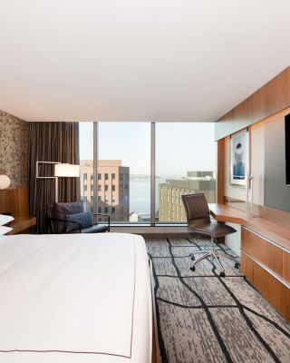 A modern hotel room features a large bed, desk, chair, wall-mounted TV, and a window with a view of city buildings and water.
