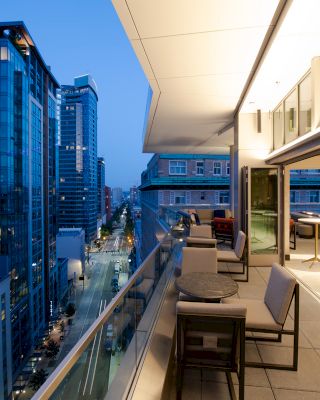 A modern balcony with seating and tables overlooks a cityscape featuring tall buildings and a street below, illuminated in the evening.
