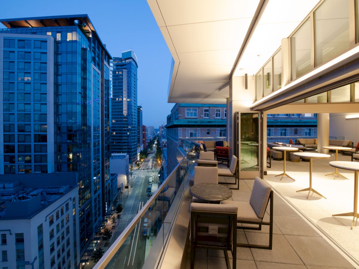 A modern cityscape view from a balcony at dusk, with seating areas and tables overlooking a street with tall buildings and a serene evening sky.