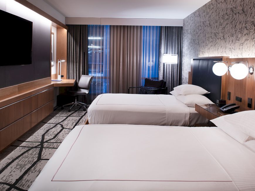 The image shows a modern hotel room with two double beds, a wall-mounted TV, a work desk, and stylish lighting and decor.