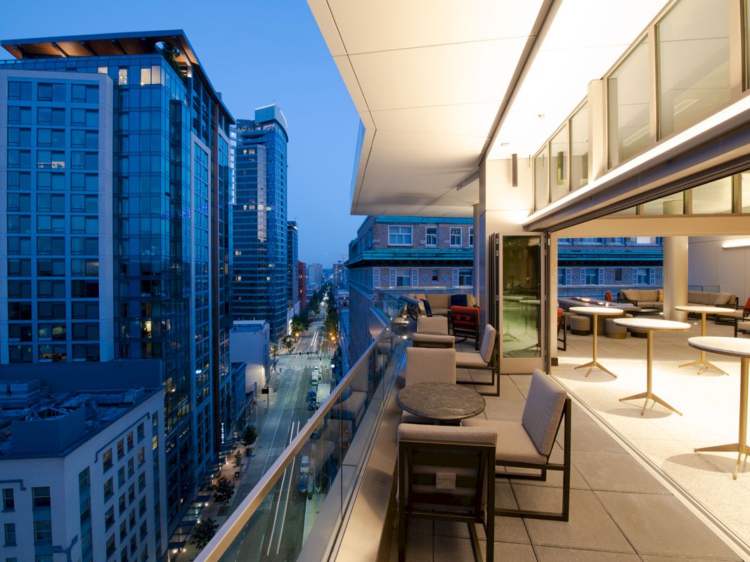A modern balcony at dusk with tables and chairs, overlooking a cityscape of tall buildings and streets illuminated by streetlights.