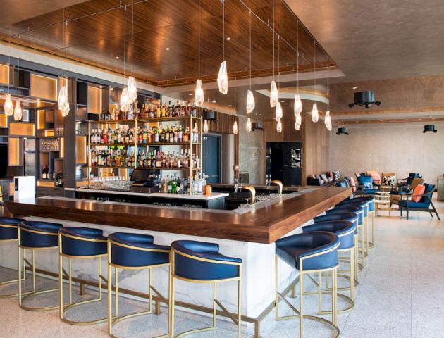 The image shows a modern bar with a wooden counter, blue and gold barstools, hanging light fixtures, and a fully stocked bar area.