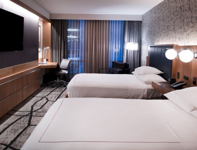 The image shows a modern hotel room with two neatly made beds, a desk with a chair, a large television, and stylish decor.