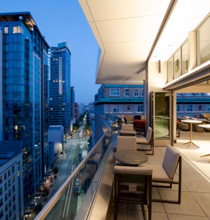 A modern balcony with tables and chairs overlooks a cityscape featuring high-rise buildings and a street at dusk.