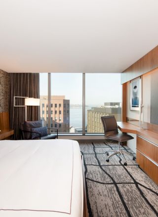 A modern hotel room with a large bed, a desk with a chair, a wall-mounted TV, and a view of buildings and water through the window.