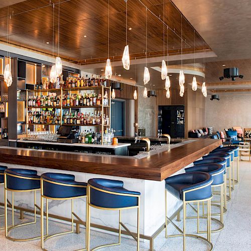 A modern bar with a wooden counter, blue cushioned bar stools, hanging lights, and a well-stocked display of bottles behind the counter.