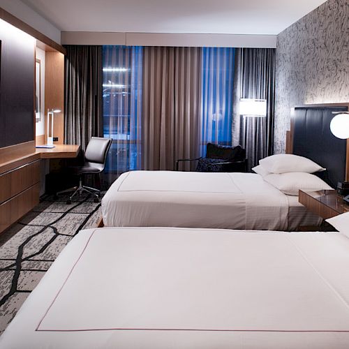 The image shows a modern hotel room with two beds, a TV, a work desk, chairs, and large windows with curtains, and a patterned wall.