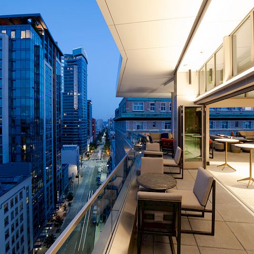 A modern balcony with seating overlooks a city street, surrounded by tall buildings, during the evening. The balcony has tables and chairs.