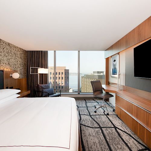 This image features a modern hotel room with a large bed, desk, chair, flat-screen TV, and a window with a view of buildings and water.