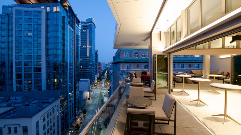 The image shows a modern cityscape view from a high-rise balcony with outdoor seating and tables, overlooking a street with tall buildings.