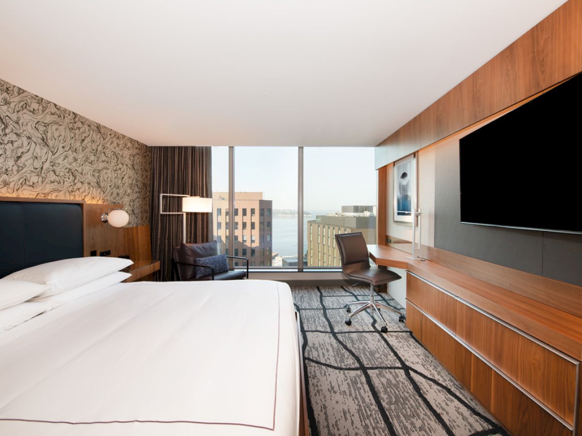 A modern hotel room with a large bed, a desk with a chair, television mounted on a wooden panel, and a cityscape view from the window.