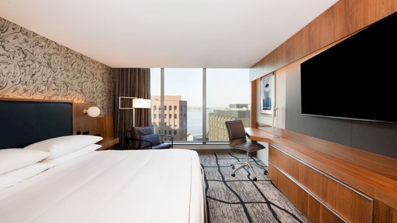 A modern hotel room with a large bed, a desk with a chair, television mounted on a wooden panel, and a cityscape view from the window.