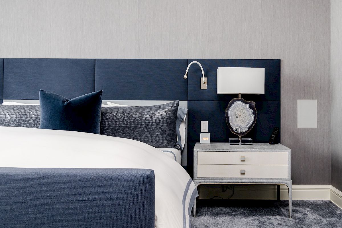 A modern bedroom features a blue upholstered bed, white bedding, a nightstand with a lamp and decorative item, and a plush carpet.