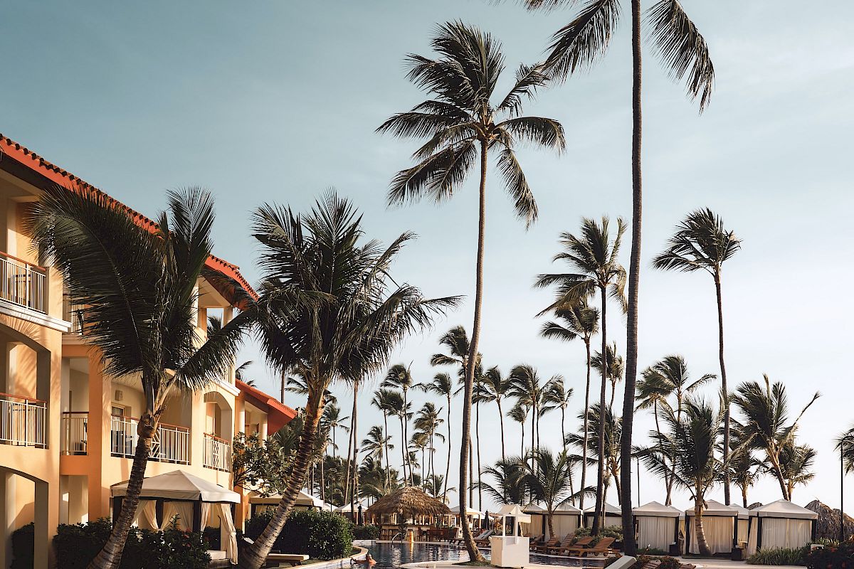 The image shows a tropical resort with palm trees, a swimming pool, lounge chairs, and a building with balconies under a clear sky.