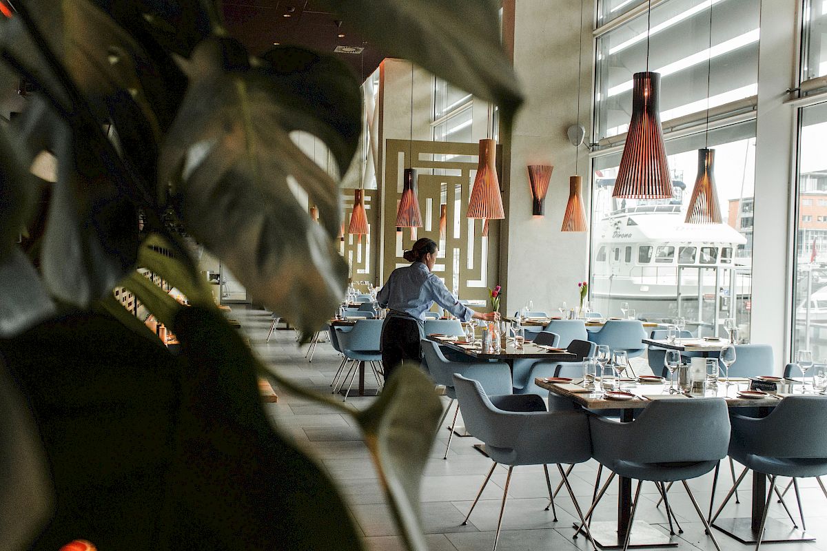 A person is setting up tables in a modern, well-lit restaurant with large plants and orange pendant lights hanging from the ceiling.