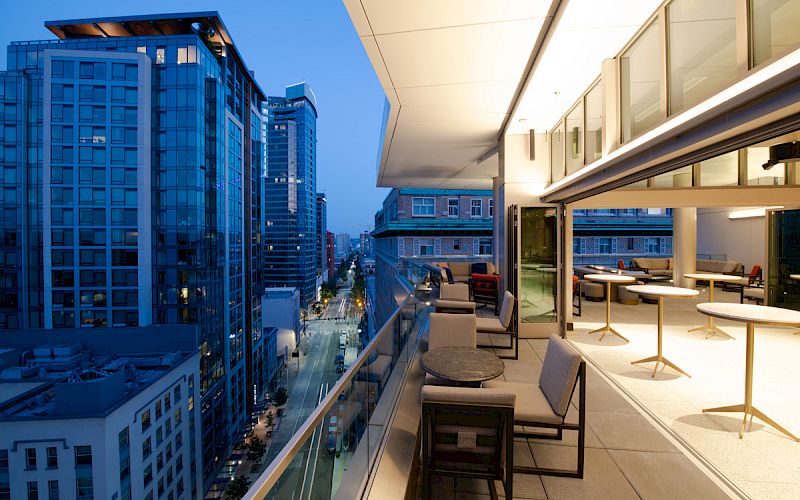 A modern rooftop terrace with outdoor seating overlooks a city street with tall buildings during the evening, showcasing an urban skyline.