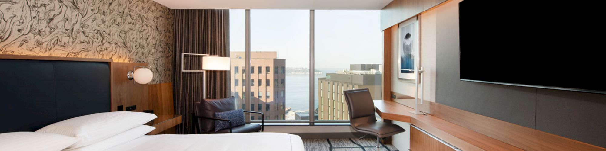A modern hotel room with a large bed, flat-screen TV, desk, chair, and a window with a city view. The decor is stylish and contemporary.
