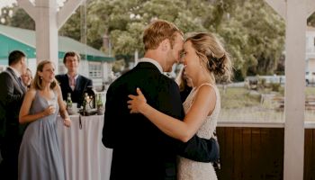 A couple is dancing closely at what appears to be a wedding, with onlookers in the background. They are surrounded by a picturesque outdoor setting.