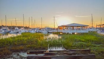 A serene marina at sunset featuring boats docked by a pier and a white pavilion, surrounded by calm waters and lush green marshland.