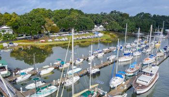 A marina with numerous sailboats and yachts docked along piers, surrounded by lush green trees and houses in the background, under a cloudy sky.