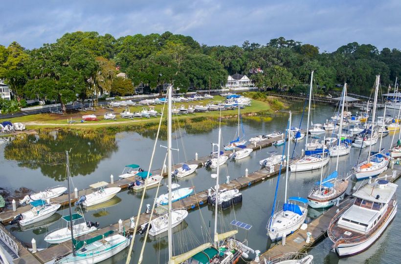 A marina with numerous sailboats and yachts docked along piers, surrounded by lush green trees and houses in the background, under a cloudy sky.