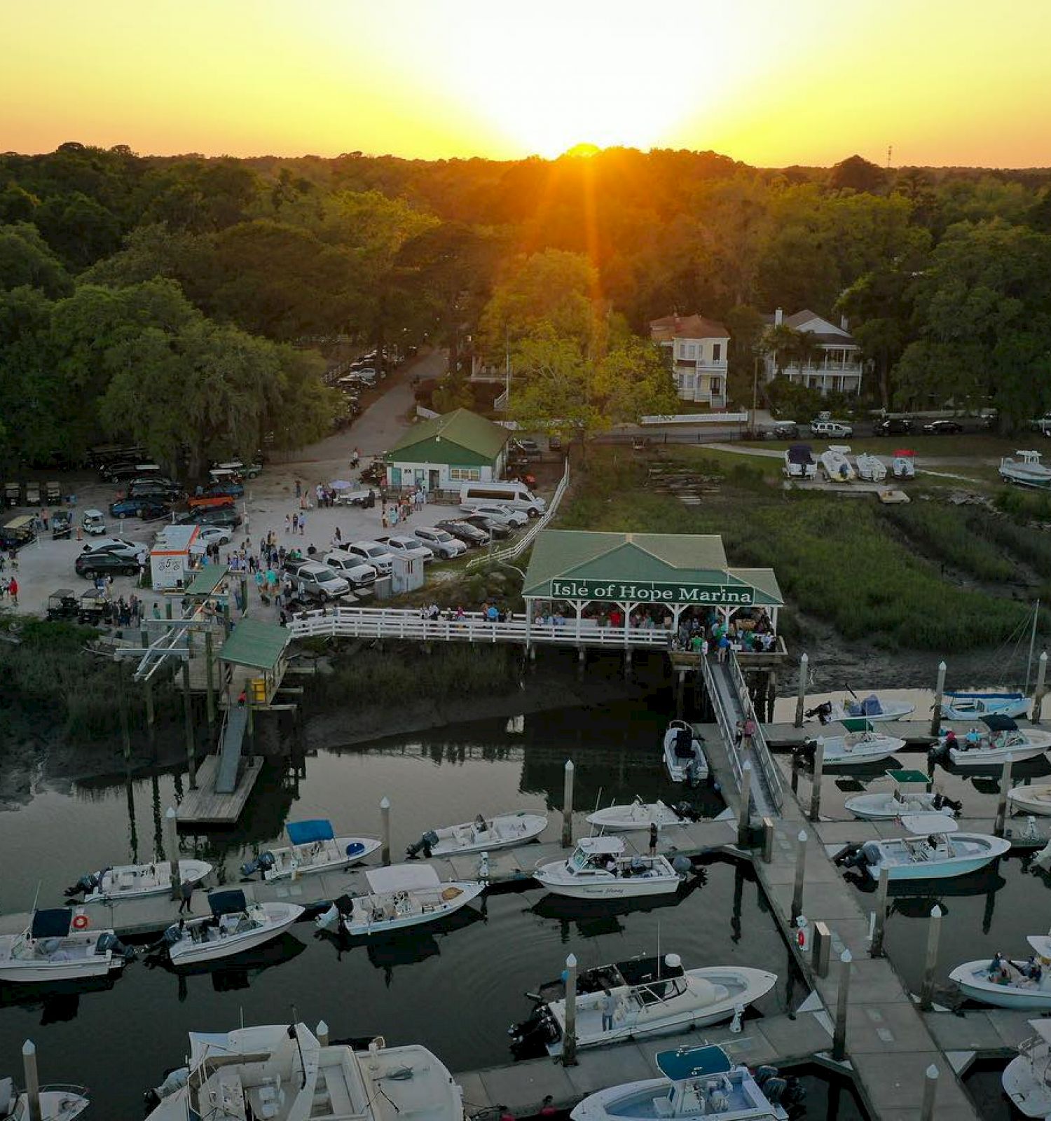 A marina at sunset with numerous boats docked, surrounded by greenery and a few buildings in the background.