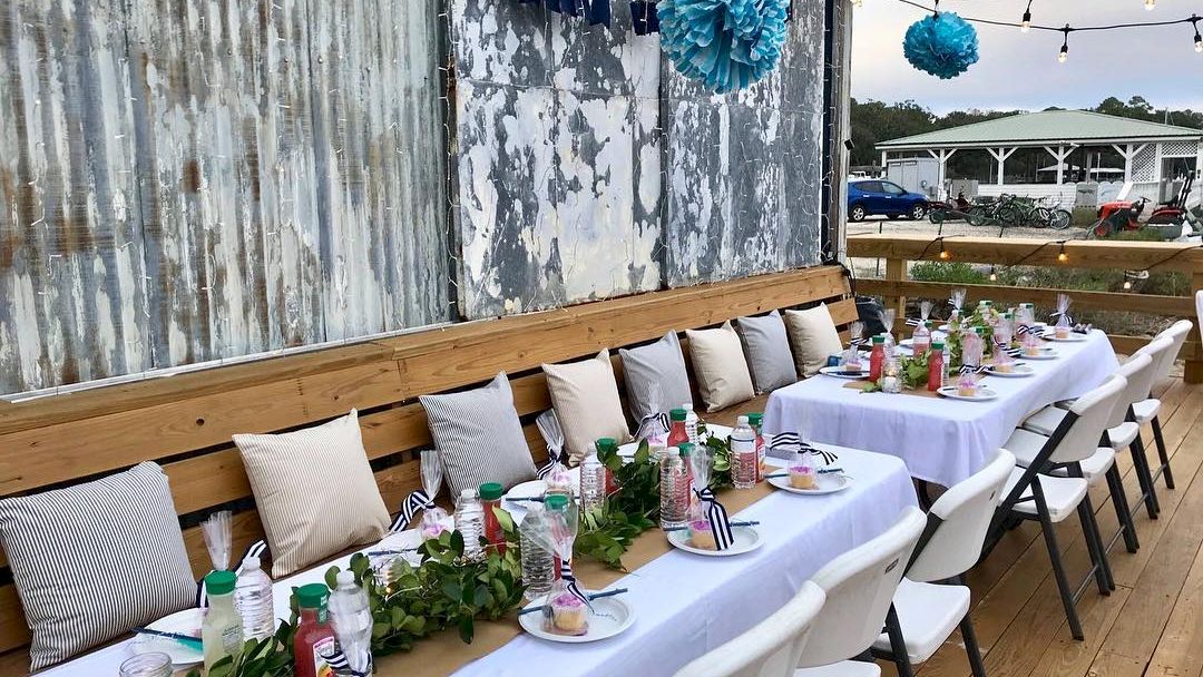 A rustic outdoor dining setup with two long tables, white tablecloths, mismatched chairs, decorative greenery, and blue paper pom-poms overhead.