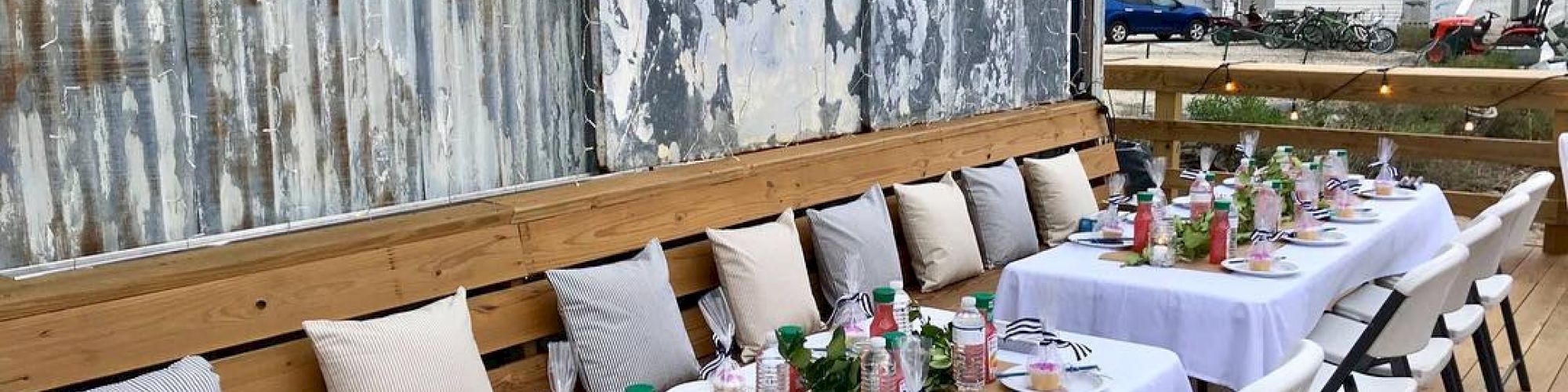 A rustic outdoor dining setup with two long tables, white tablecloths, mismatched chairs, decorative greenery, and blue paper pom-poms overhead.