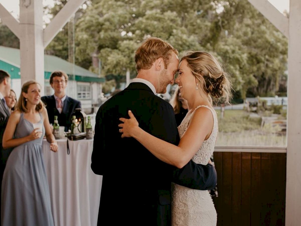 A couple shares a close dance at an outdoor event while guests in formal attire watch and mingle nearby, with a green building visible in the background.