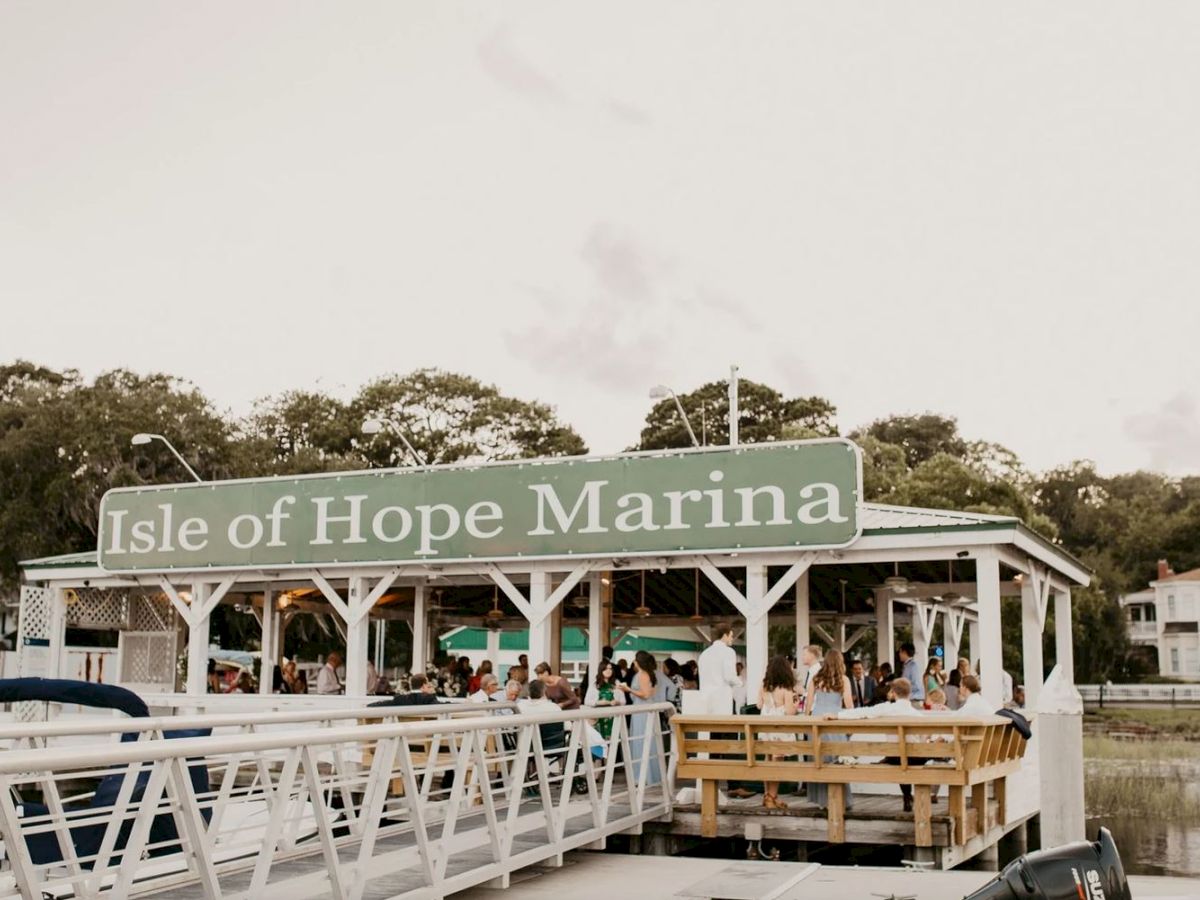 The image shows the Isle of Hope Marina with a gathering of people under a pavilion near the water, surrounded by trees and docks.